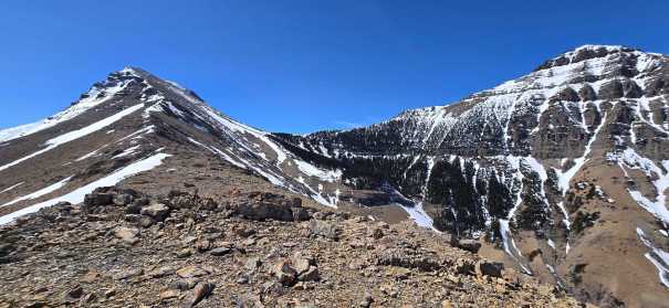 Panoramic view of rocky mountain peaks with patches of snow and clear blue sky.