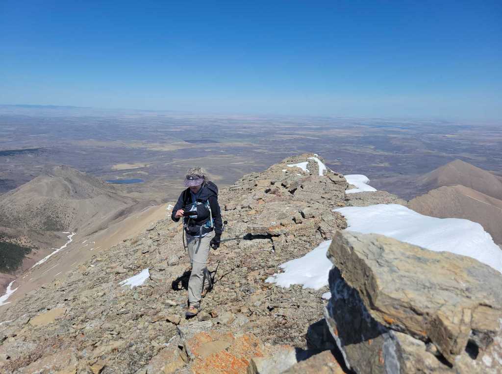 A hiker navigating rocky terrain near a summit, with a panoramic view of the surrounding landscape, including patches of snow and distant hills under a clear blue sky.