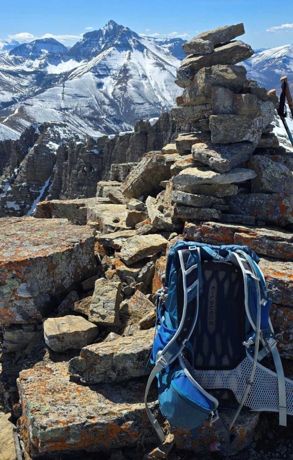 Summit view featuring a stone cairn and a blue backpack on rocky terrain, with snowy mountains in the background.