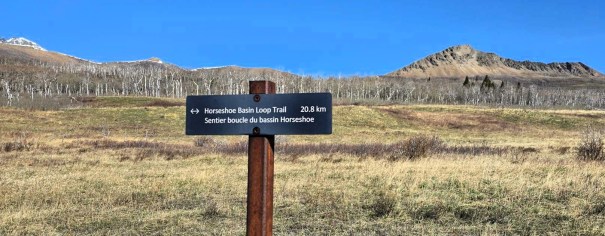 Trail sign indicating the 'Horseshoe Basin Loop Trail' with a distance of 20.8 km, set against a backdrop of grassy fields and distant mountains under a clear blue sky.