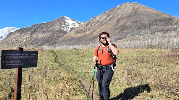 A hiker standing on a trail near a signpost indicating the Horseshoe Basin Loop Trail, with mountains in the background and snow-capped peaks visible.