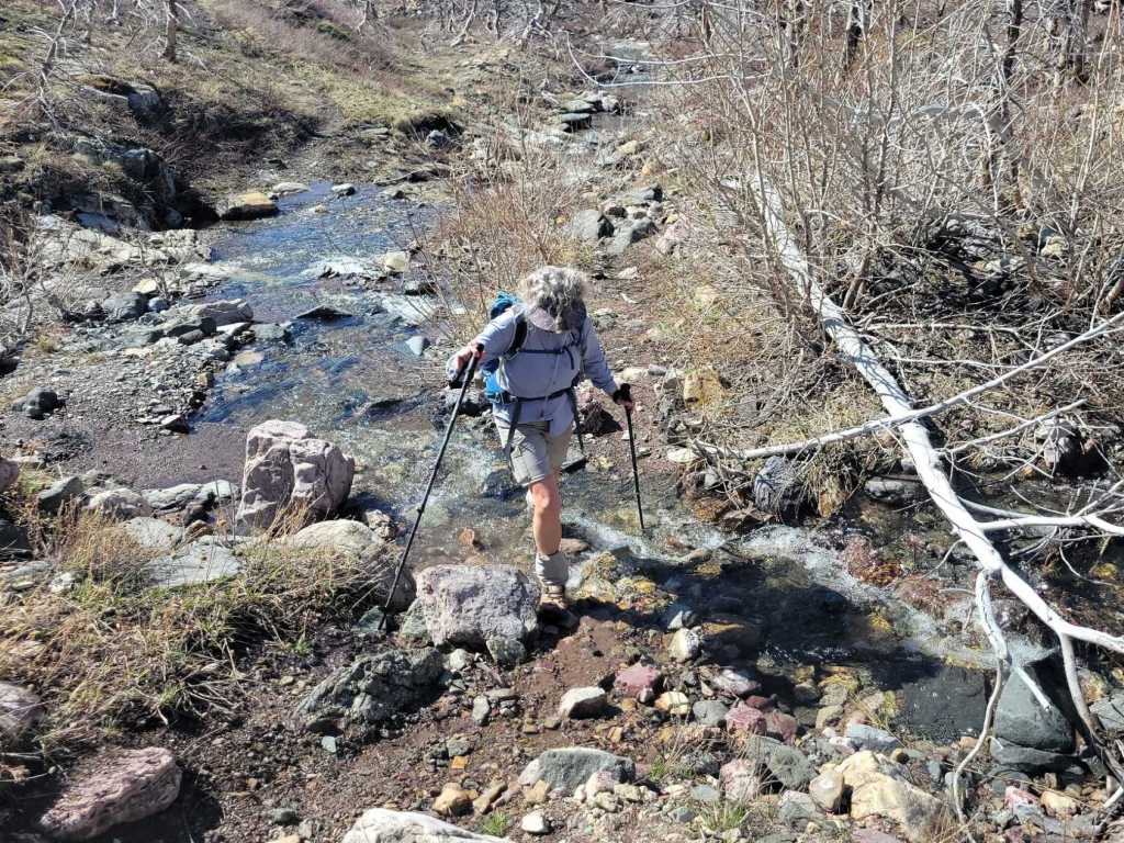 A hiker navigating through a rocky creek bed, using trekking poles for support. The surrounding area features dry grass, scattered stones, and bare tree branches.