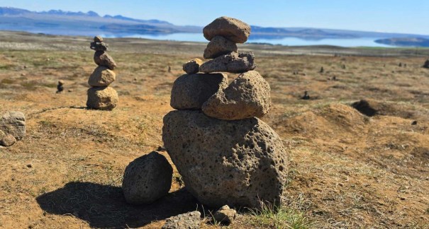 A stone cairn made of stacked volcanic rocks on a grassy, outdoor landscape, with a lake and mountains in the background under a clear blue sky.