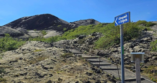 Pathway leading up to Grábrók Crater with a directional sign, surrounded by moss-covered terrain and lush greenery under a clear blue sky.