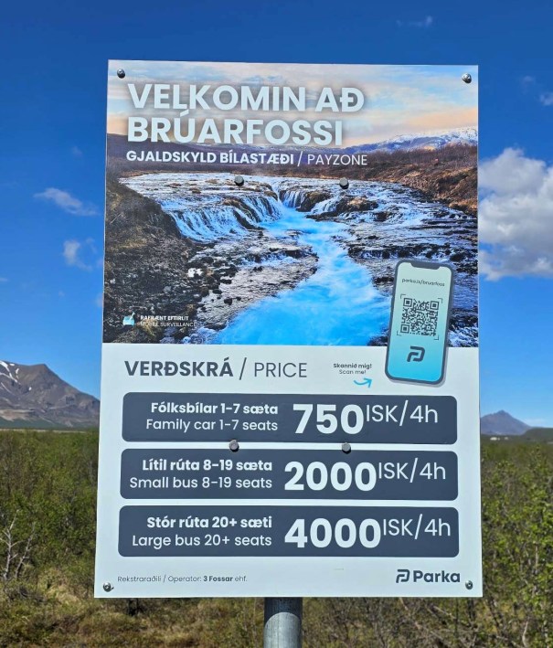 Sign welcoming visitors to Bruarfoss with pricing details for different vehicle types, featuring a backdrop of the waterfall and blue waters.