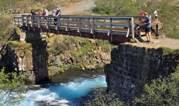 A wooden bridge spans over a vibrant blue river with people standing on it, surrounded by lush greenery and rocky terrain.
