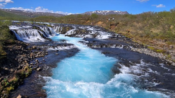 A stunning view of Bruarfoss Waterfall in Iceland, featuring bright ice blue water cascading over rocky ledges into a glacial river, surrounded by lush greenery and distant mountains under a clear blue sky.
