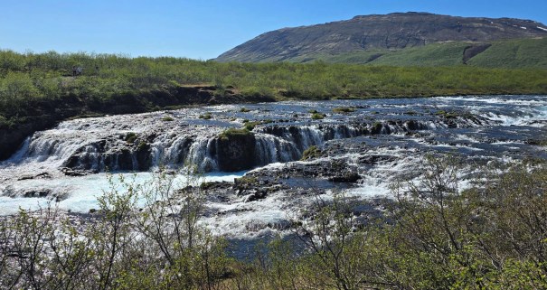 A picturesque view of Bruarfoss Waterfall, featuring cascading ice blue water flowing over rocky ledges, surrounded by lush greenery and a distant mountain in the background.