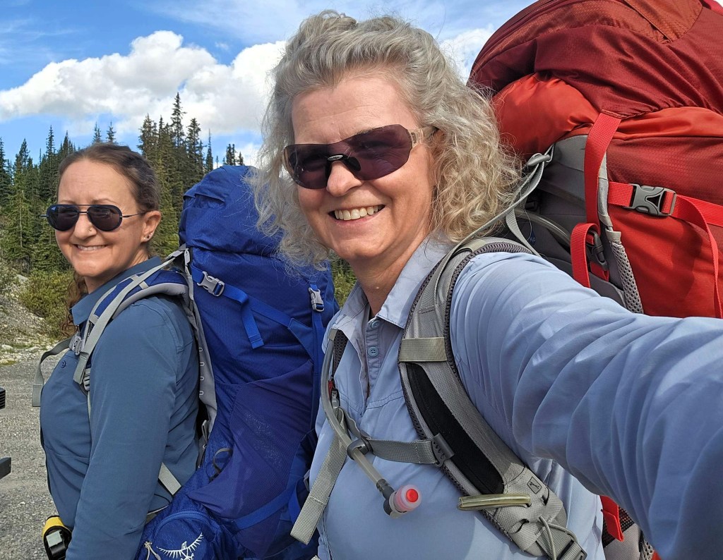 Two women smiling while wearing hiking gear and carrying backpacks, with a scenic background of trees and clouds.