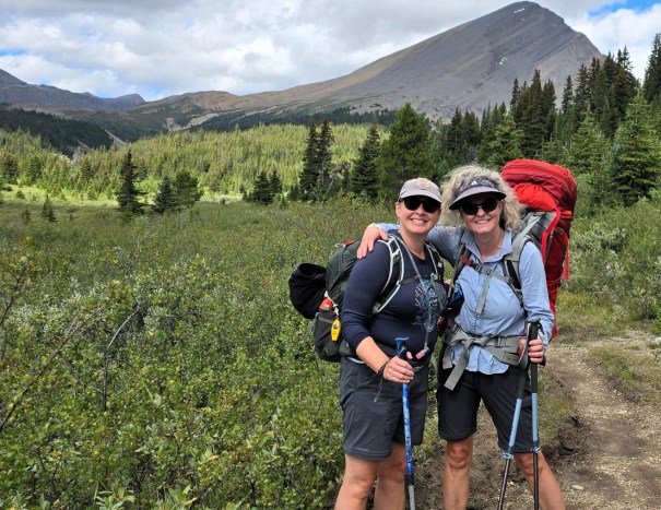 Two women with backpacks posing for a photo on a hiking trail surrounded by lush greenery and mountains in the background.