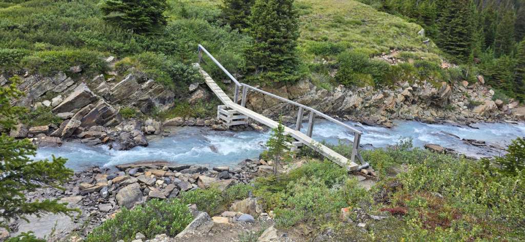 A wooden footbridge crossing a swift river, surrounded by rocky terrain and lush greenery.