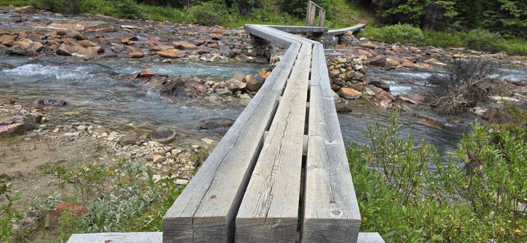 A wooden footbridge crossing a clear stream, surrounded by rocks and greenery.