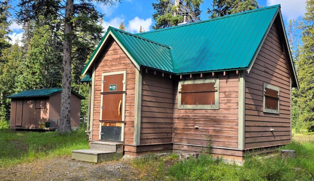 A wooden warden's cabin with a green metal roof, featuring closed windows and a wooden door, situated in a grassy area surrounded by trees.