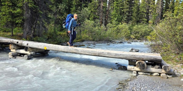 A hiker crossing a rustic wooden bridge over a river, wearing a large backpack and using hiking poles, surrounded by dense greenery and trees.