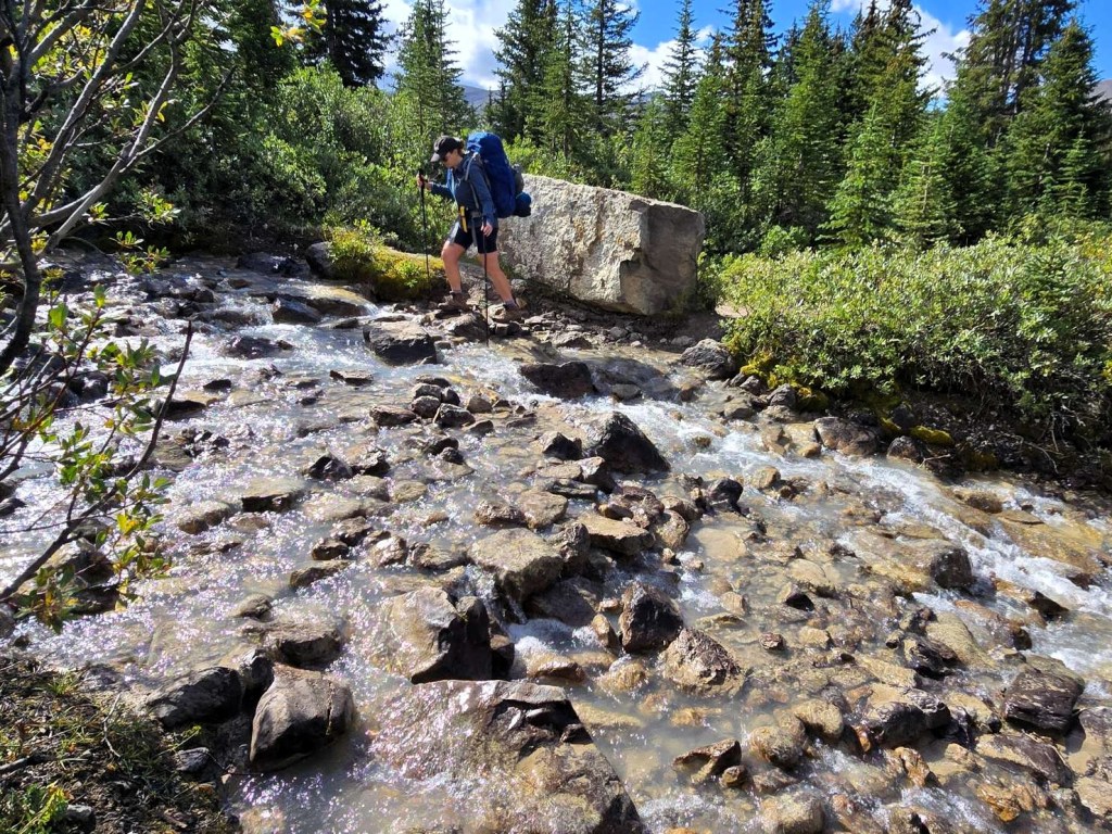 A hiker navigating a rocky stream bed with a large boulder nearby, surrounded by lush greenery and tall trees.