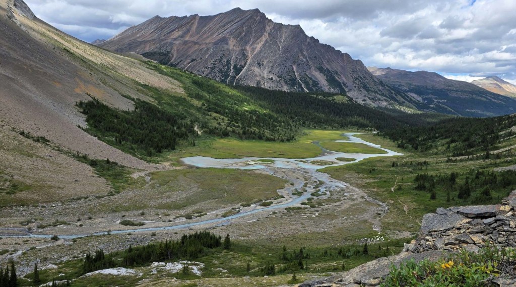A scenic view of a green valley with a winding river, mountains in the background, and a partly cloudy sky.