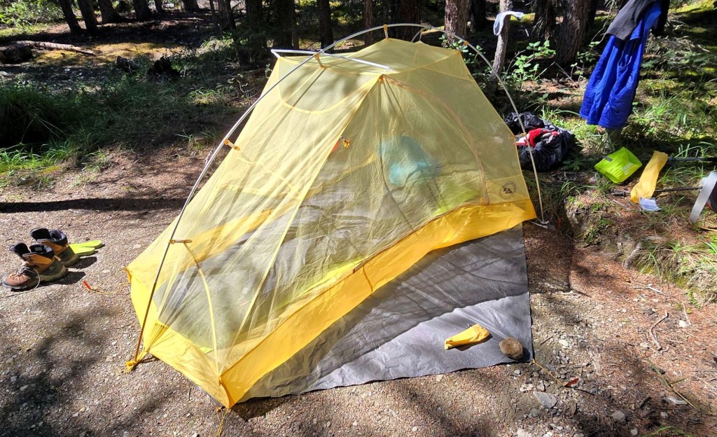 A lightweight two-person tent set up in a forested campsite, with hiking boots and gear placed nearby.