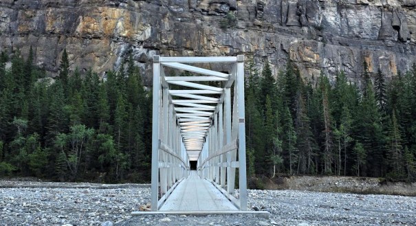 A white metal bridge leading into a wooded area with cliffs in the background.