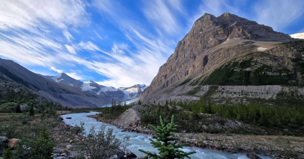 A scenic view of a river winding through a valley, surrounded by majestic mountains and a clear blue sky with wispy clouds.