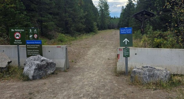 Entrance to South Castle Valley Trail showing signs for no vehicles and registration information, surrounded by trees and rocky barriers.