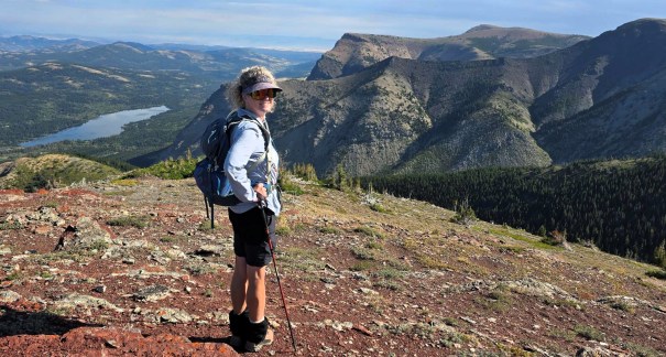 A hiker standing on a rocky mountain trail, looking over a vast landscape of hills and a lake in the distance.