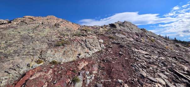 Rocky terrain leading up to a summit with patches of soil and sparse vegetation under a blue sky.