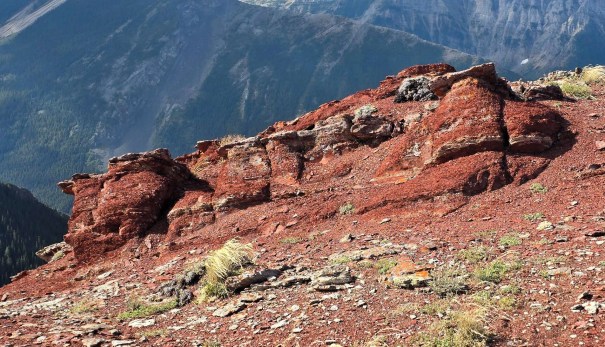 Rocky terrain with reddish-brown rock formations and sparse vegetation under a mountainous backdrop.