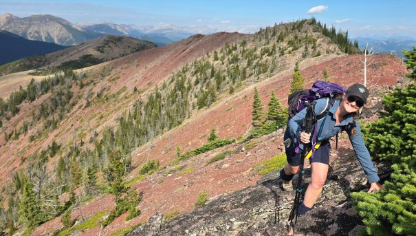 A hiker ascending a rocky trail on a mountainous landscape, surrounded by greenery and distant mountains under a clear sky.