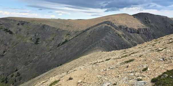 A panoramic view of rugged mountain ridges with rocky terrain and patches of greenery under a cloudy sky.