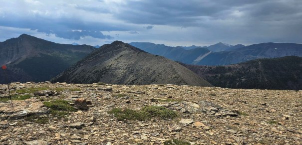 A panoramic view of rocky mountain peaks under a cloudy sky, featuring a rugged landscape and a gentle slope in the foreground.