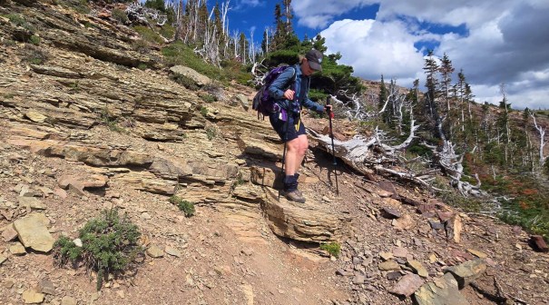 A hiker carefully descending a rocky slope while using trekking poles, surrounded by a rugged landscape with sparse trees and blue sky.