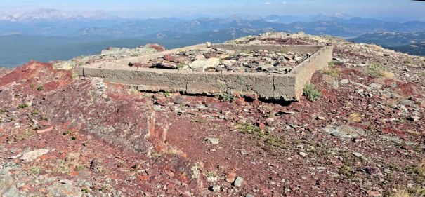 Concrete foundation remains on a rocky mountaintop, surrounded by a panoramic view of distant mountains and valleys.