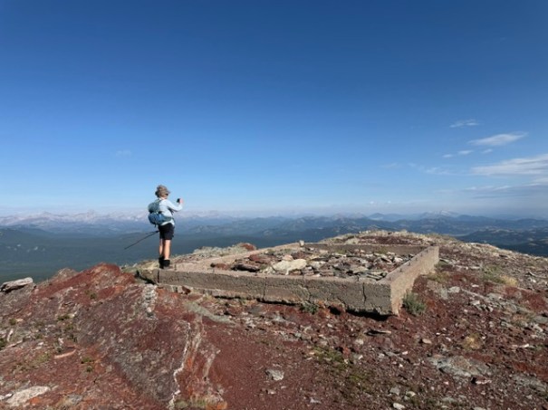 A hiker standing on a rocky mountain summit, taking a photo of the landscape, with a stone foundation visible in the foreground and distant mountains in the background.