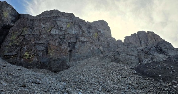 A rocky landscape featuring steep cliffs and a gravelly slope under a cloudy sky.