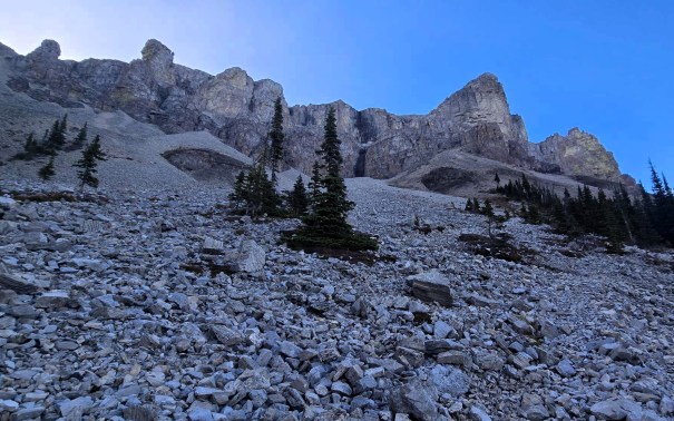 A rocky hillside with scattered boulders and a few trees, leading up to steep rocky cliffs under a clear blue sky.