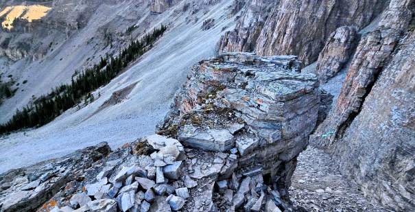 A rocky cliff with layered stone formations, overlooking a sloped landscape with sparse vegetation and distant mountains.