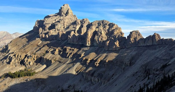 A majestic mountain peak with rugged cliffs under a blue sky, showcasing geological formations and shadows on the slopes.