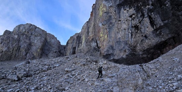 A hiker ascending a rocky slope in a mountainous area, with towering cliffs in the background and a clear blue sky.