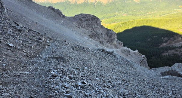 A rocky mountain slope with loose gravel leading downwards, surrounded by green forested areas and distant hills in the background.