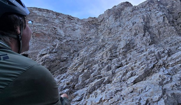 A person wearing a helmet and glasses looking up at a steep rocky cliff face.