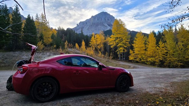 A red sports car parked on a gravel road with mountains and autumn foliage in the background.