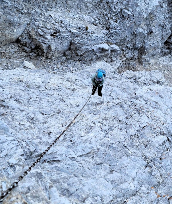 A climber ascending a rocky surface using a metal chain for support, surrounded by rugged gray rocks.