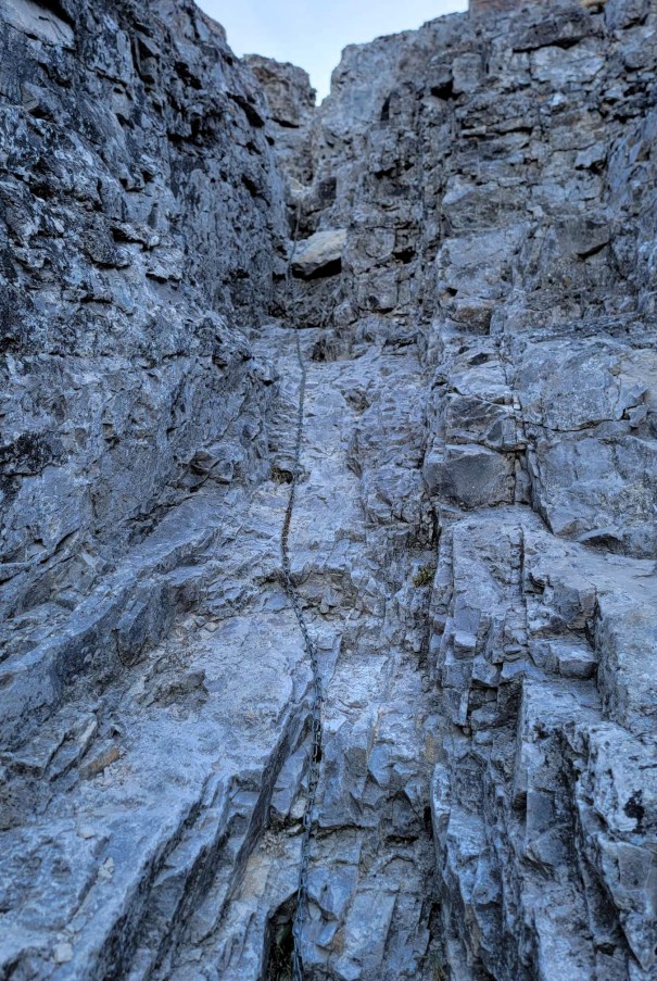 A vertical rock face with a chain bolted into the cliffside for climbing assistance, surrounded by jagged stone formations.