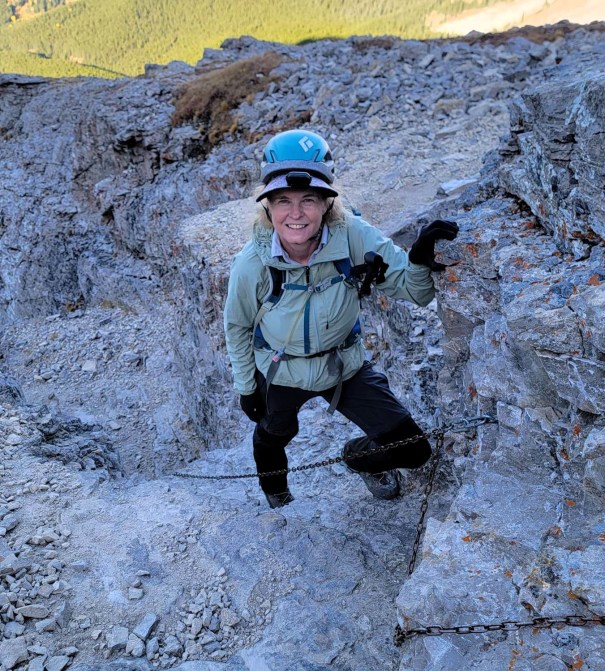 A climber smiles while navigating a rocky terrain, wearing a helmet and harness, with a mountain landscape in the background.