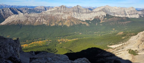 Panoramic view of rugged mountain range with lush green valley and patches of autumn foliage below.