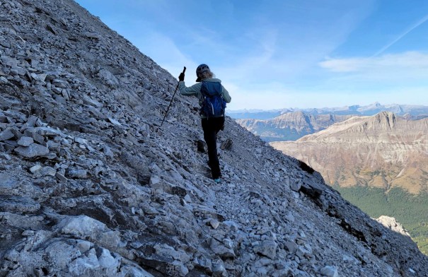 A hiker ascending a rocky mountain slope, using trekking poles, with a scenic view of distant mountains and a blue sky in the background.