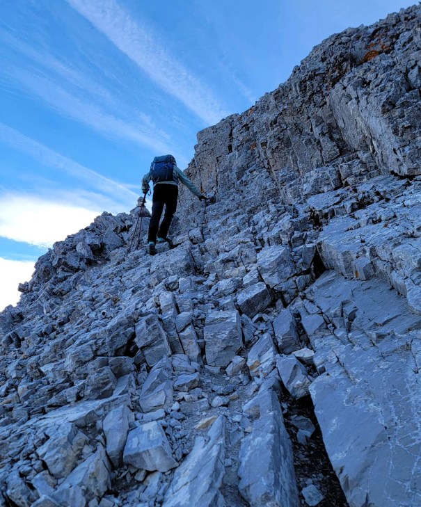 A hiker ascends a rocky mountain slope under a blue sky with wispy clouds.