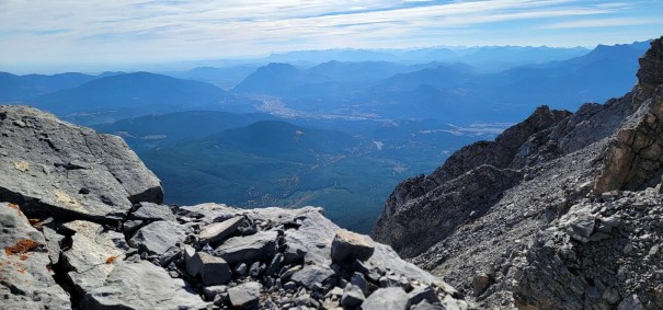 A panoramic view from a mountain summit, showcasing rocky foreground with distant mountains and a valley below under a blue sky.