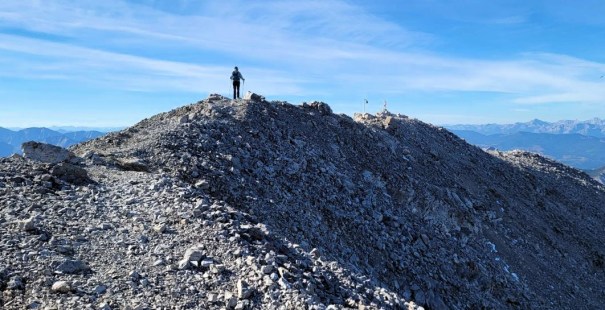 Person standing on a rocky mountain peak with a clear blue sky in the background.