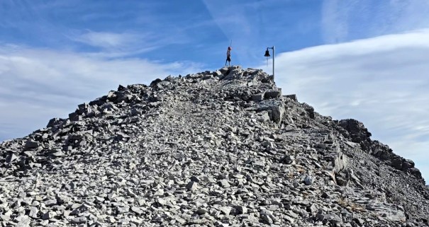 A person standing at the top of a rocky mountain peak, with a flag on a pole in the background and a cloudy blue sky above.